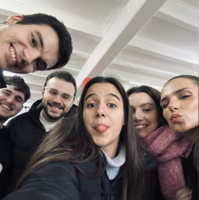 Group of six young adults smiling and posing for a selfie indoors, one woman sticking out her tongue playfully.
