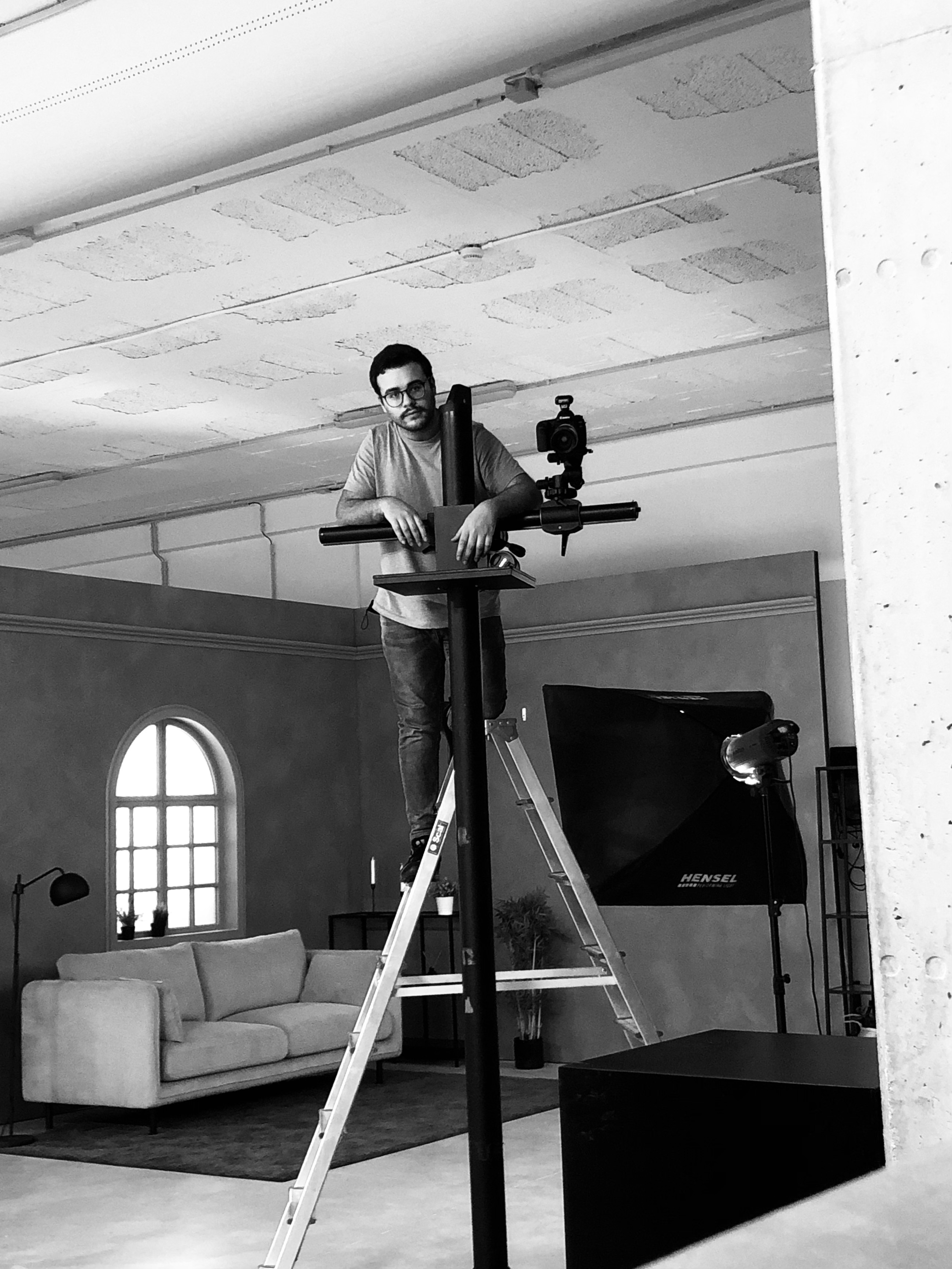 A man standing on a ladder operating a professional camera mounted on a vertical rig in a studio with a sofa, window, and studio light.