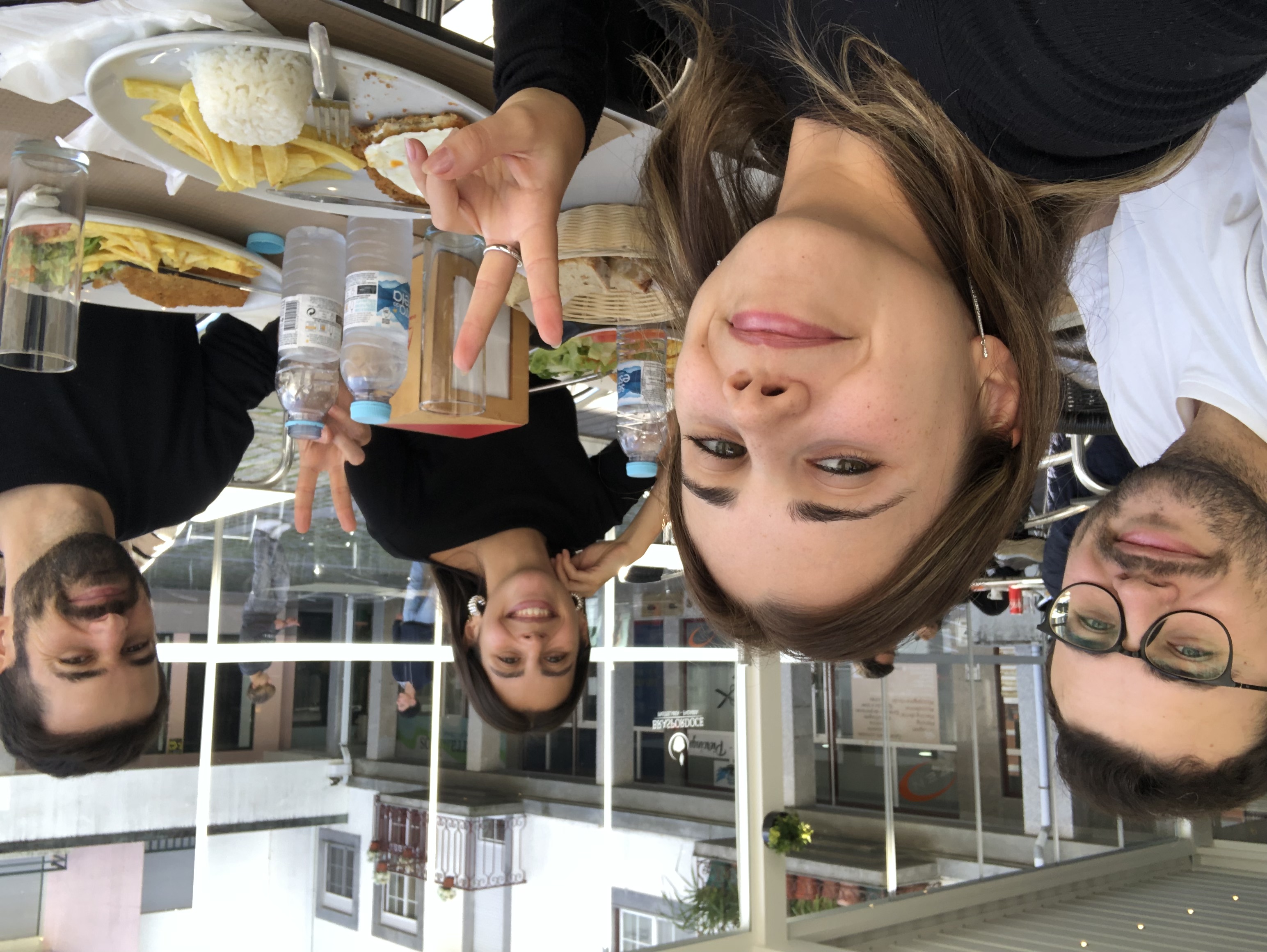 Four friends sitting at an outdoor café table enjoying a meal with plates of fries, rice, and water bottles, smiling and making peace signs.