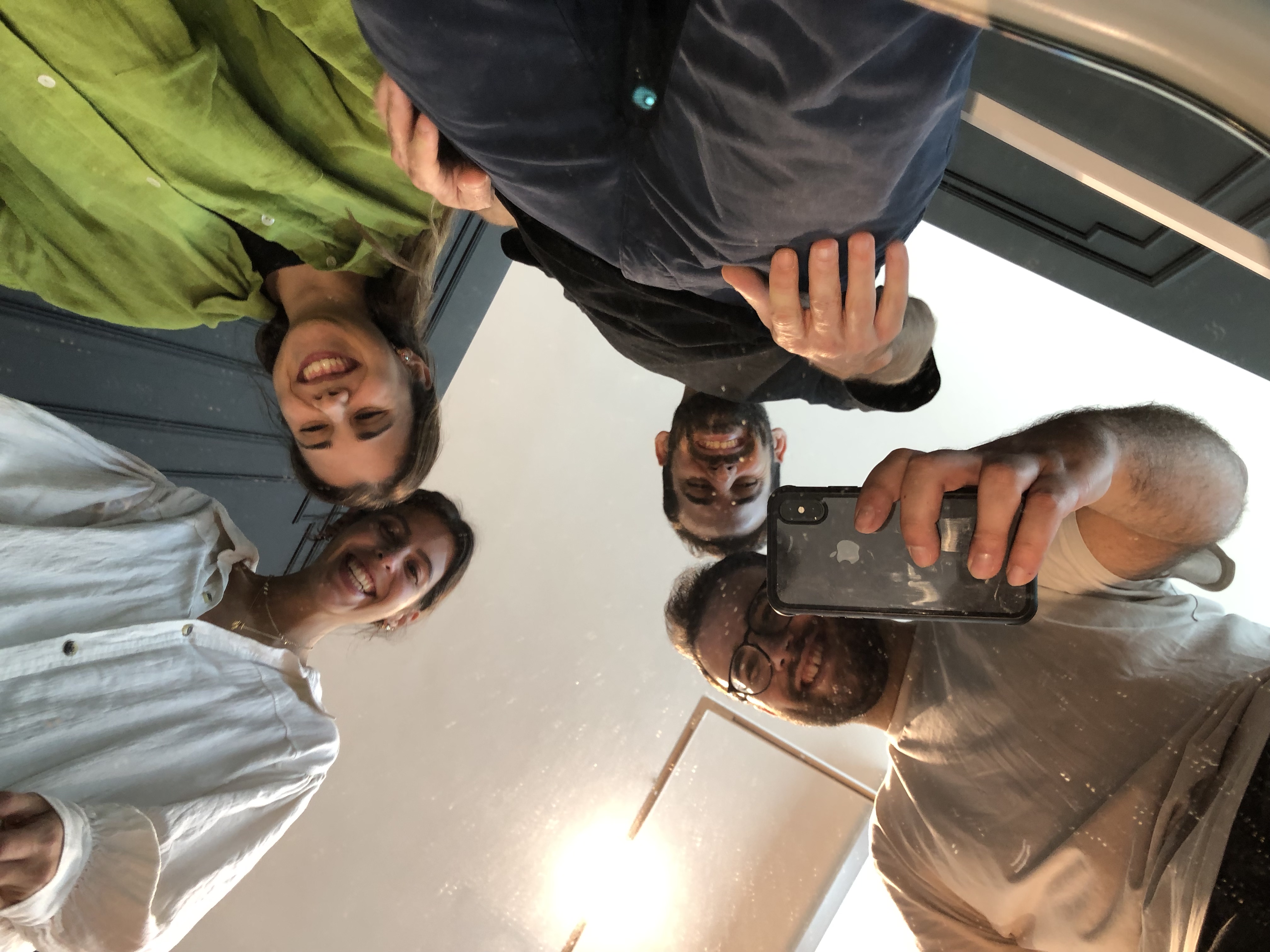 Four smiling people looking down at the camera, one holding a smartphone to take a photo.