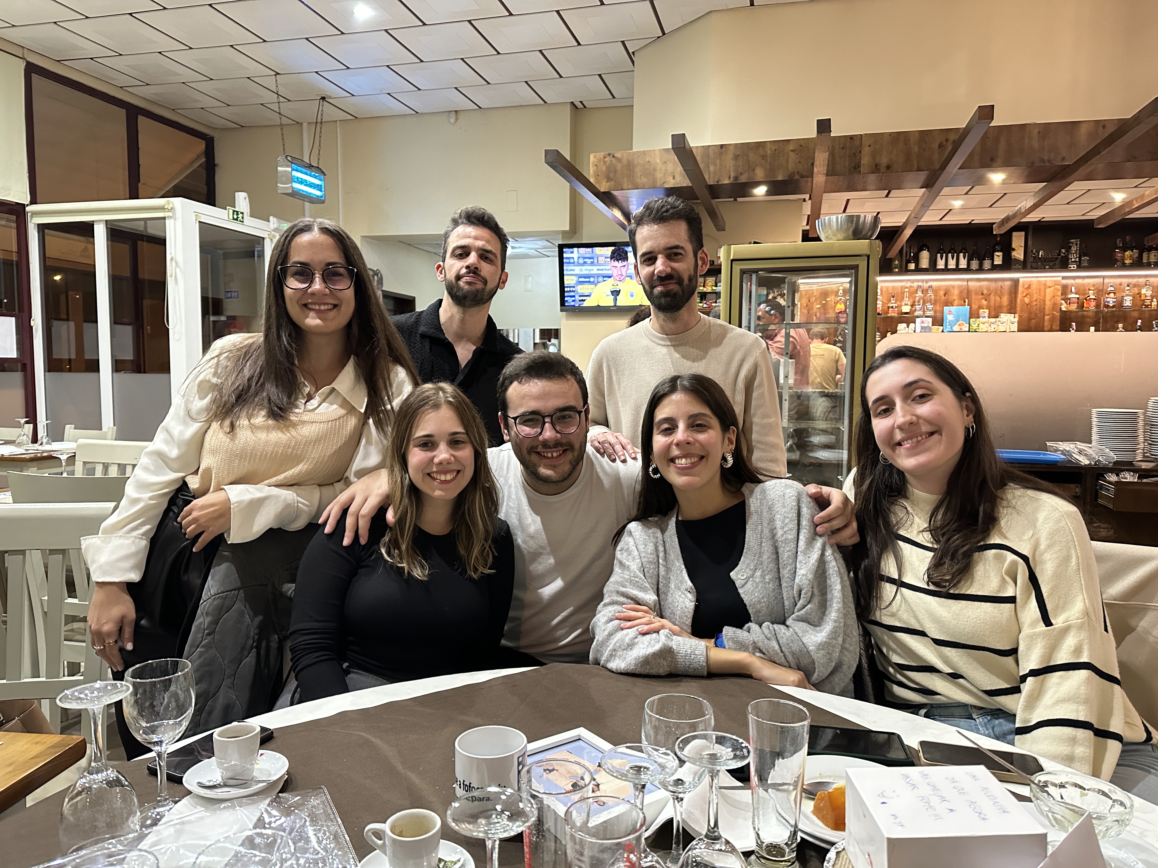 Group of seven young adults smiling and posing around a dining table with glasses, cups, and plates in a cozy restaurant setting.