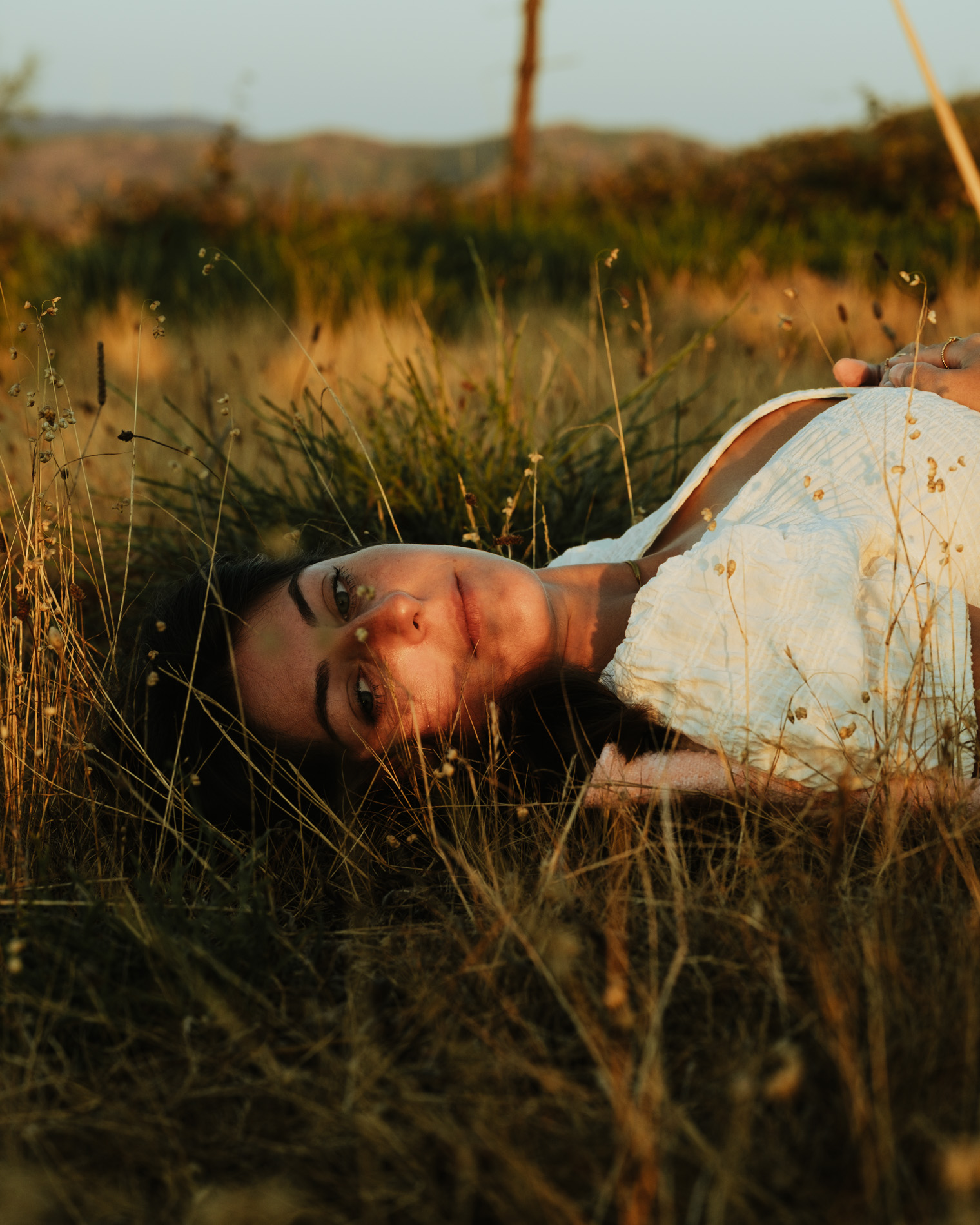 Woman lying in tall dry grass wearing a white dress, looking calmly at the camera during golden hour.