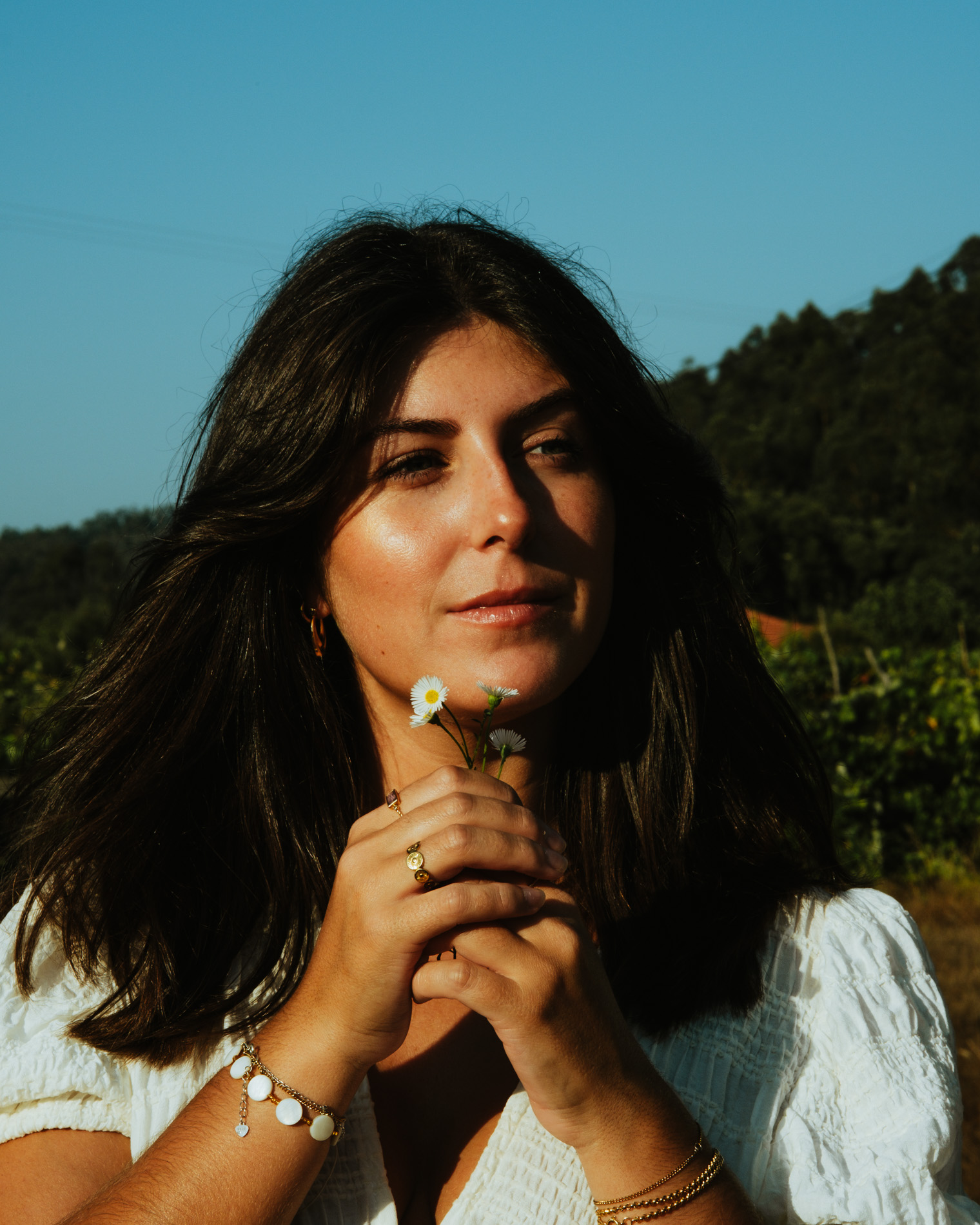 Woman with dark hair holding small white flowers near her face outdoors with a forest background.