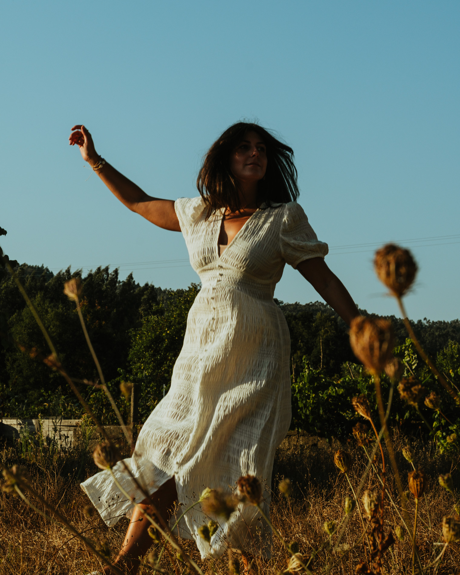 Woman in a white dress dancing in a dry grassy field with green trees and clear blue sky in the background.