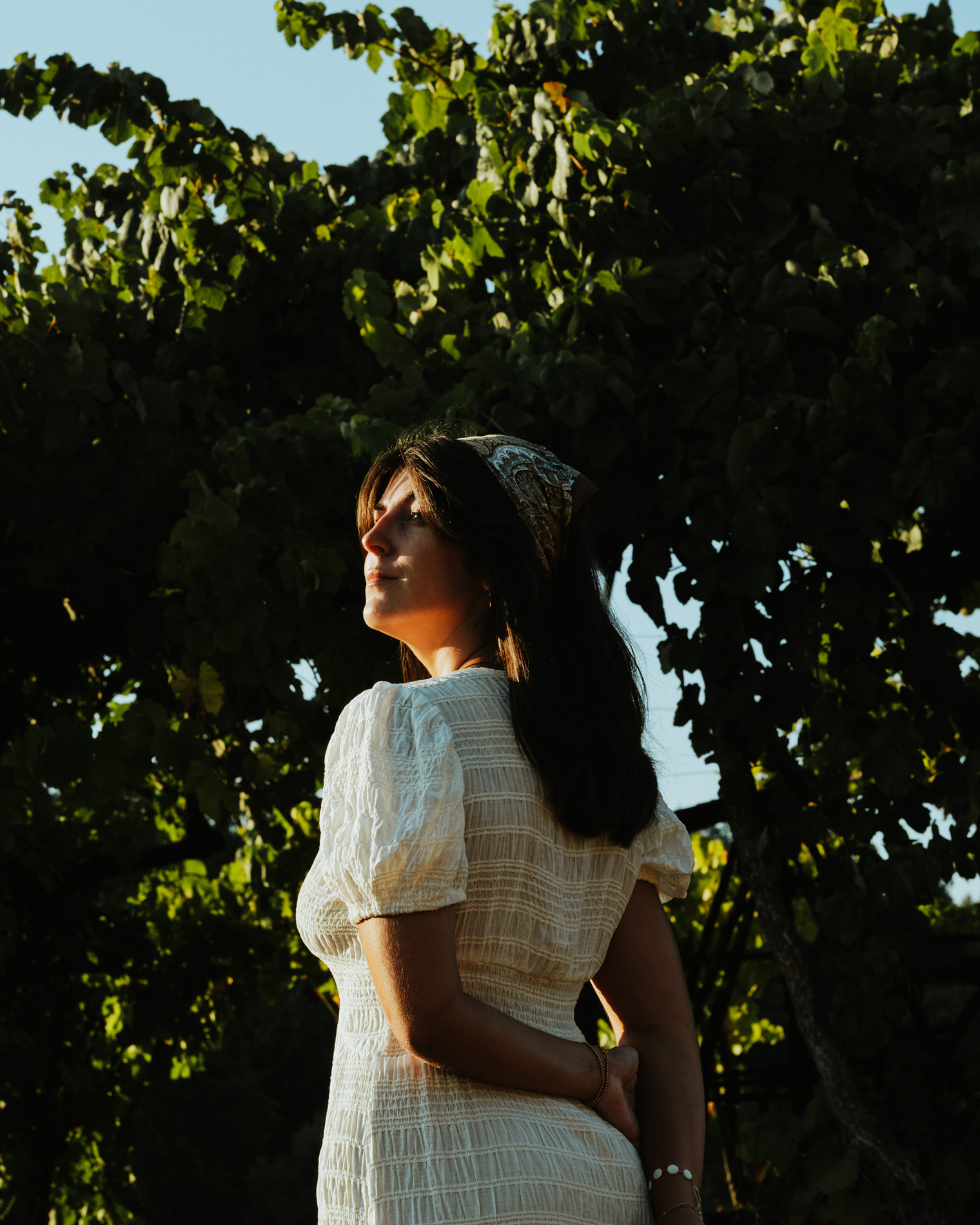 Woman in a white dress and patterned headscarf standing outdoors near green leafy trees in warm sunlight.