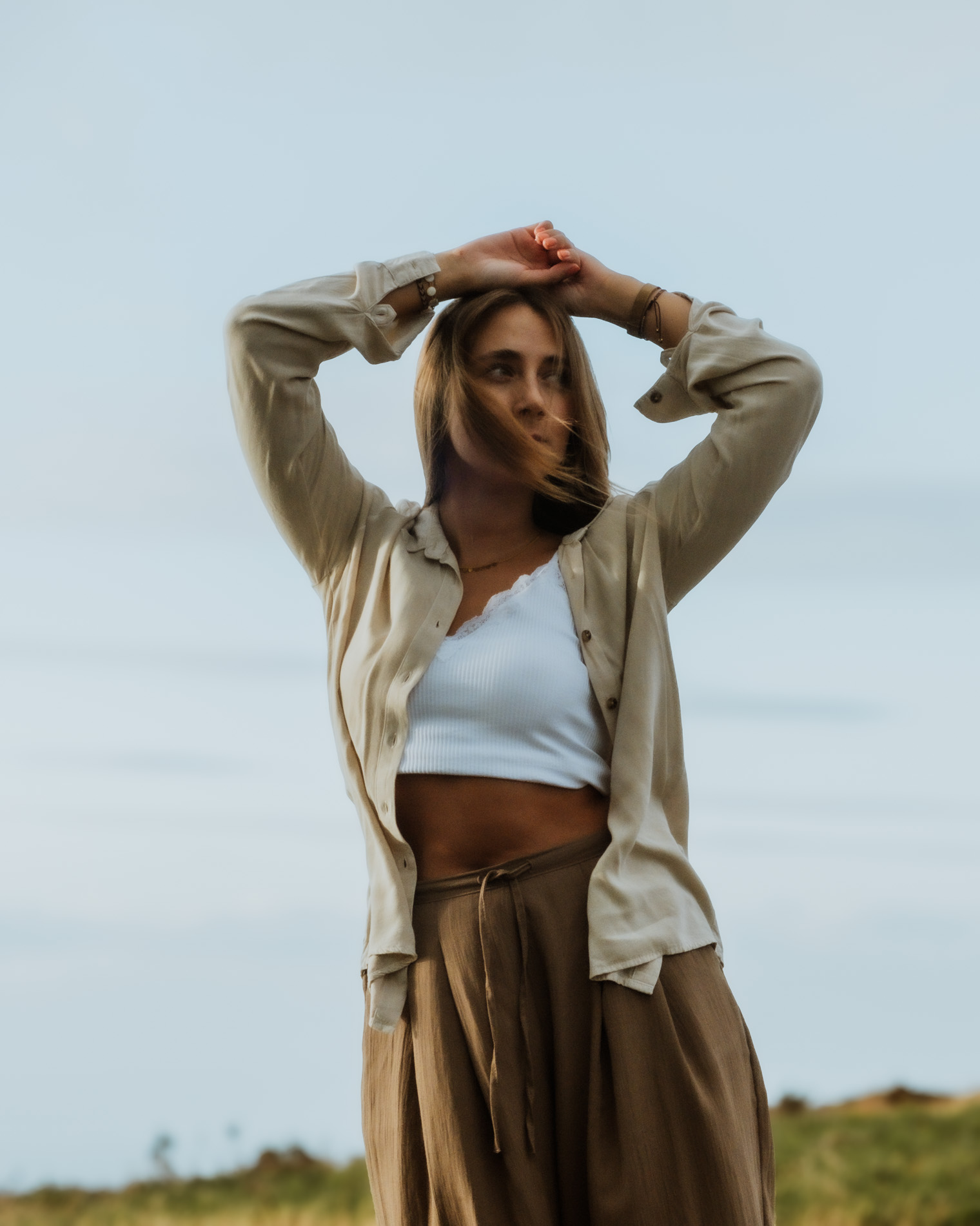 Woman in beige shirt and brown skirt standing outdoors with arms raised above her head against a cloudy sky.