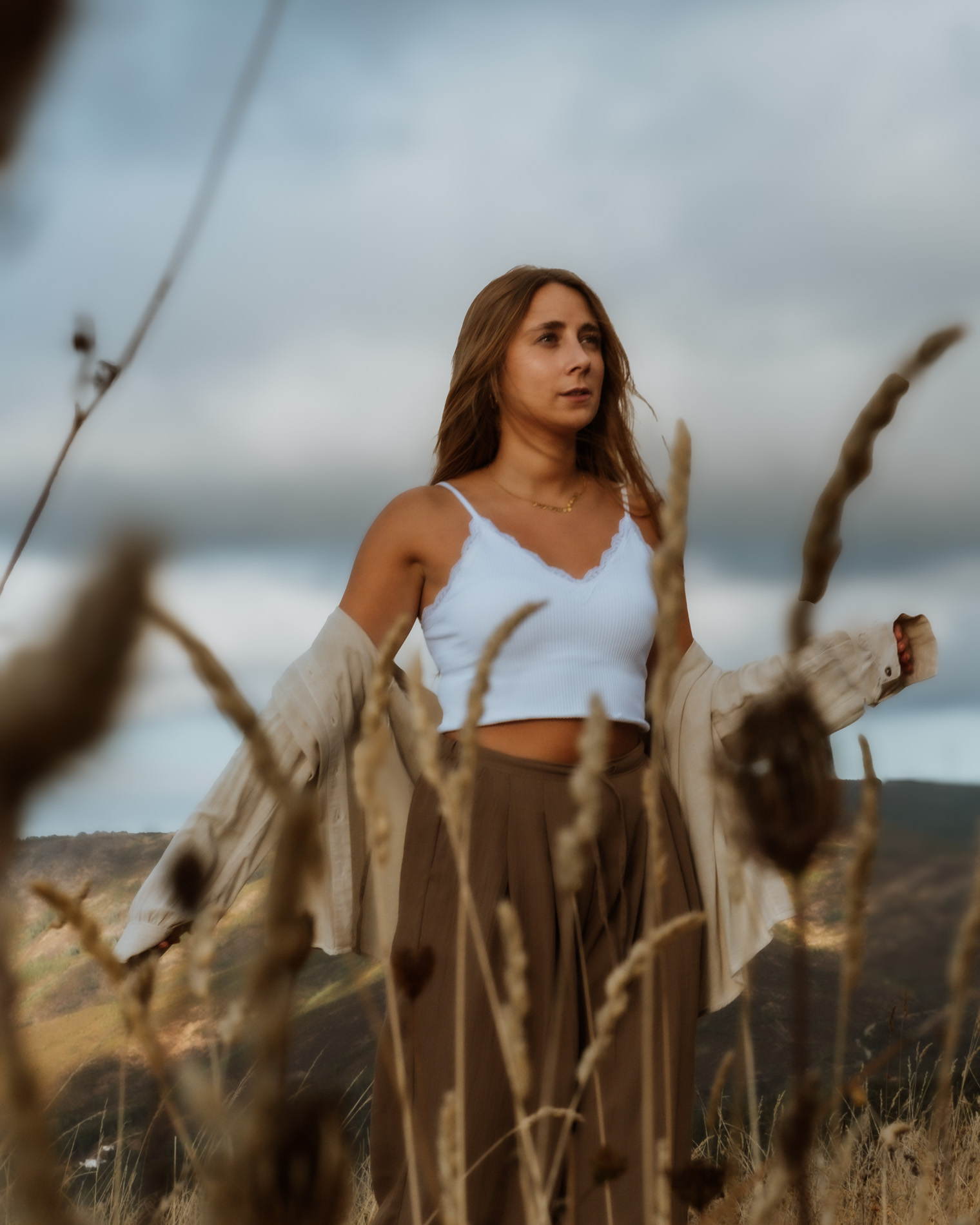 Woman with long brown hair standing outdoors in a field with tall dry grass, wearing a white crop top and brown skirt with a beige jacket draped over her arms.