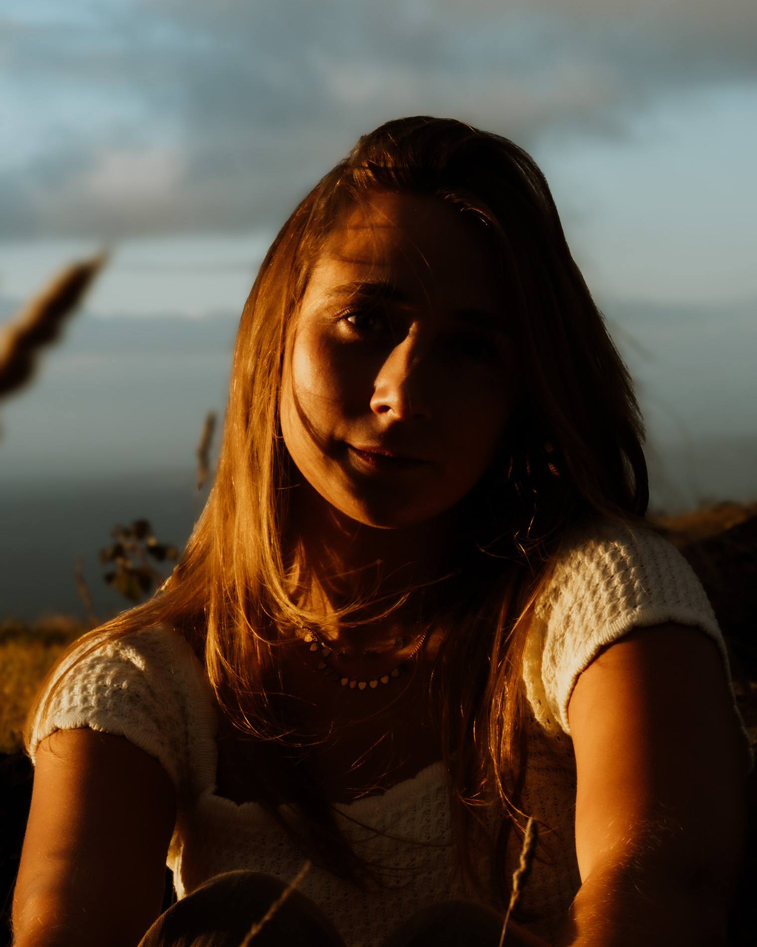 Young woman with long hair wearing a white knitted top, illuminated by warm sunset light against a blurred outdoor background.
