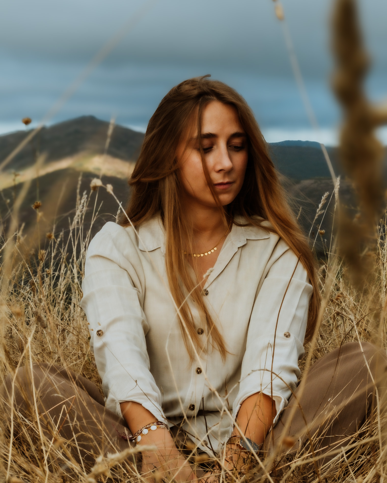 Woman with long hair wearing a light shirt and sitting in dry grass with mountains and cloudy sky in the background.