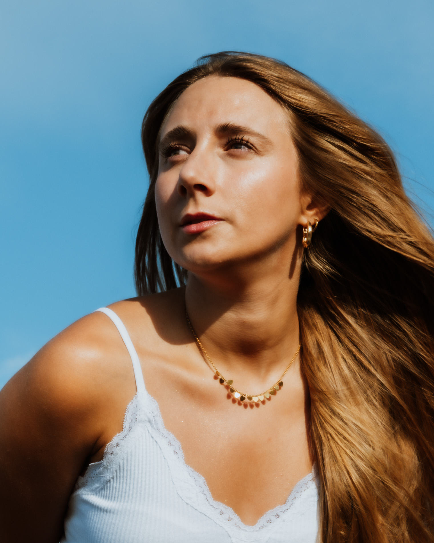 Young woman with long flowing hair wearing a white lace-trimmed tank top and gold necklace looking to the side against a blue sky.