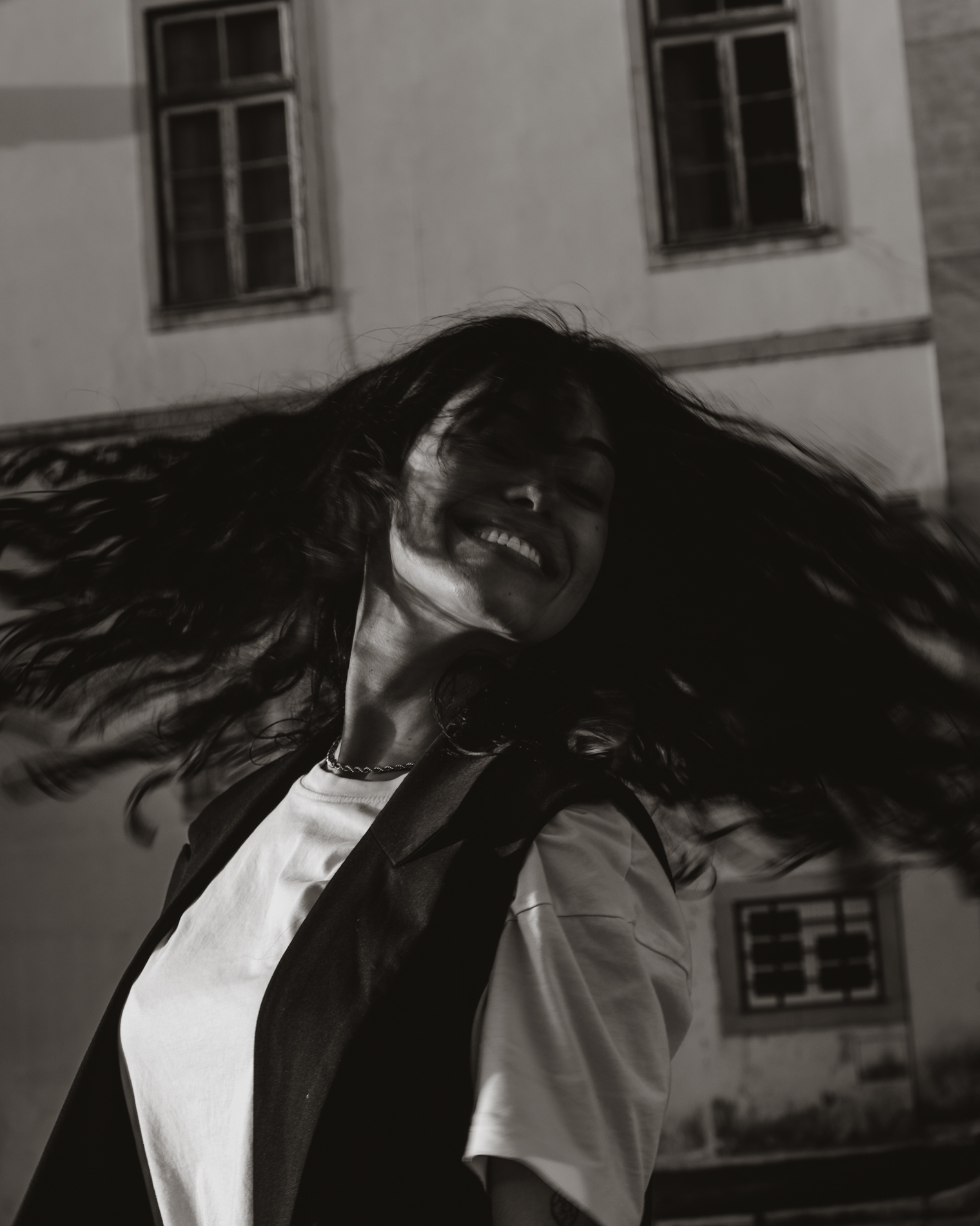 Smiling woman with long hair flipping it outdoors in front of a building with windows.