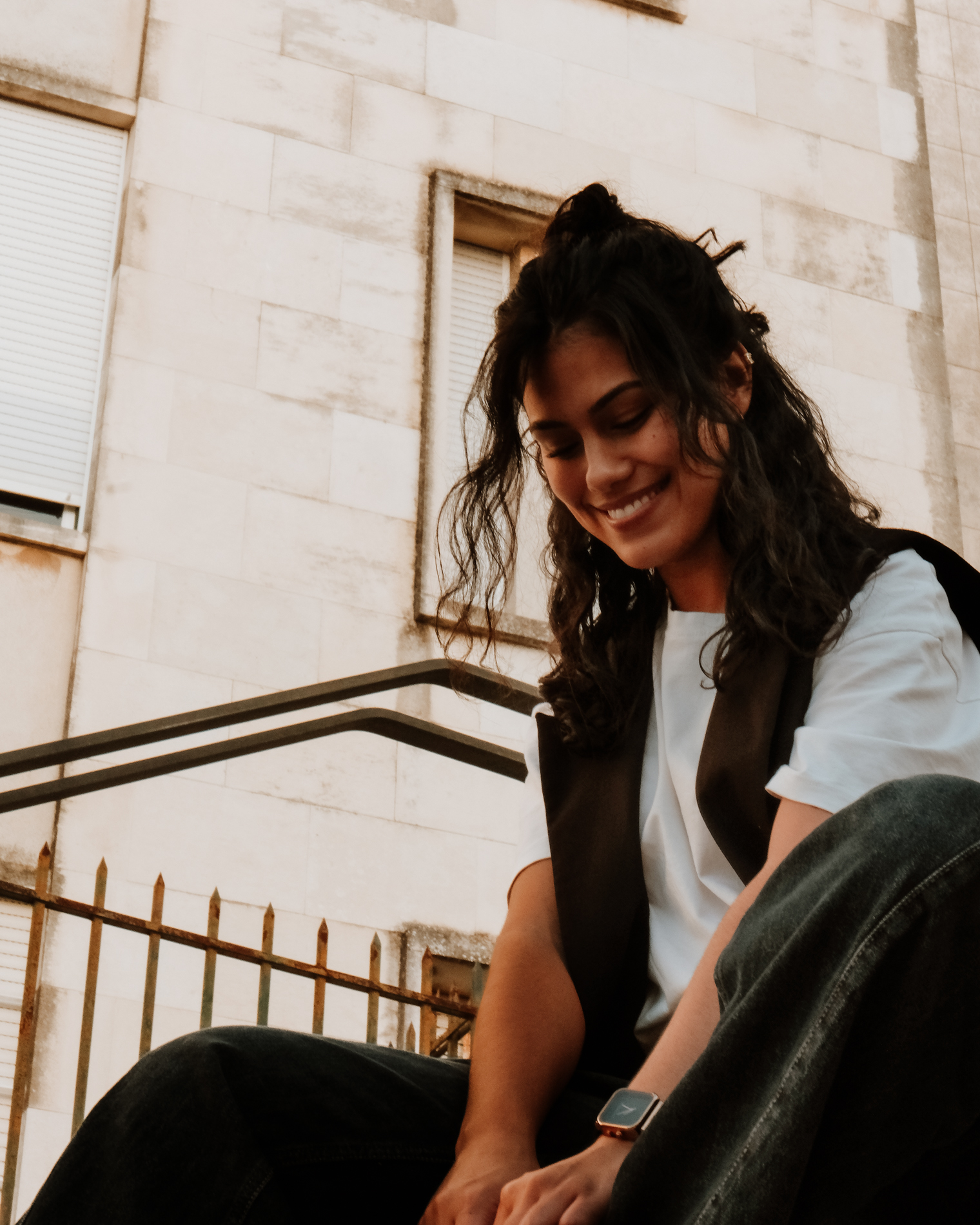 Smiling woman with curly hair sitting outdoors wearing a white t-shirt, black vest, and a smartwatch.