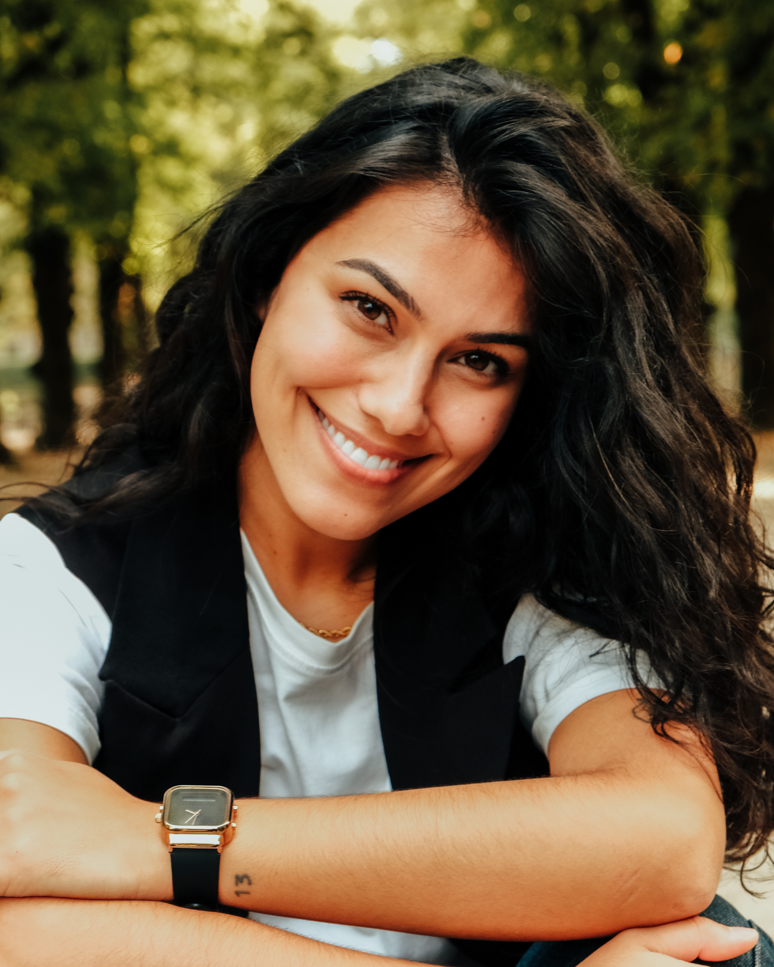 Smiling woman with dark curly hair wearing a white shirt and black vest outdoors.