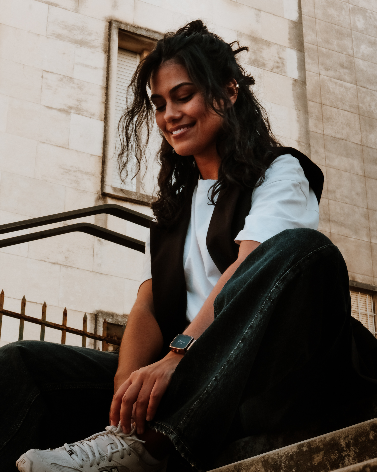 Smiling woman with curly hair sitting on outdoor steps, tying her white sneaker.