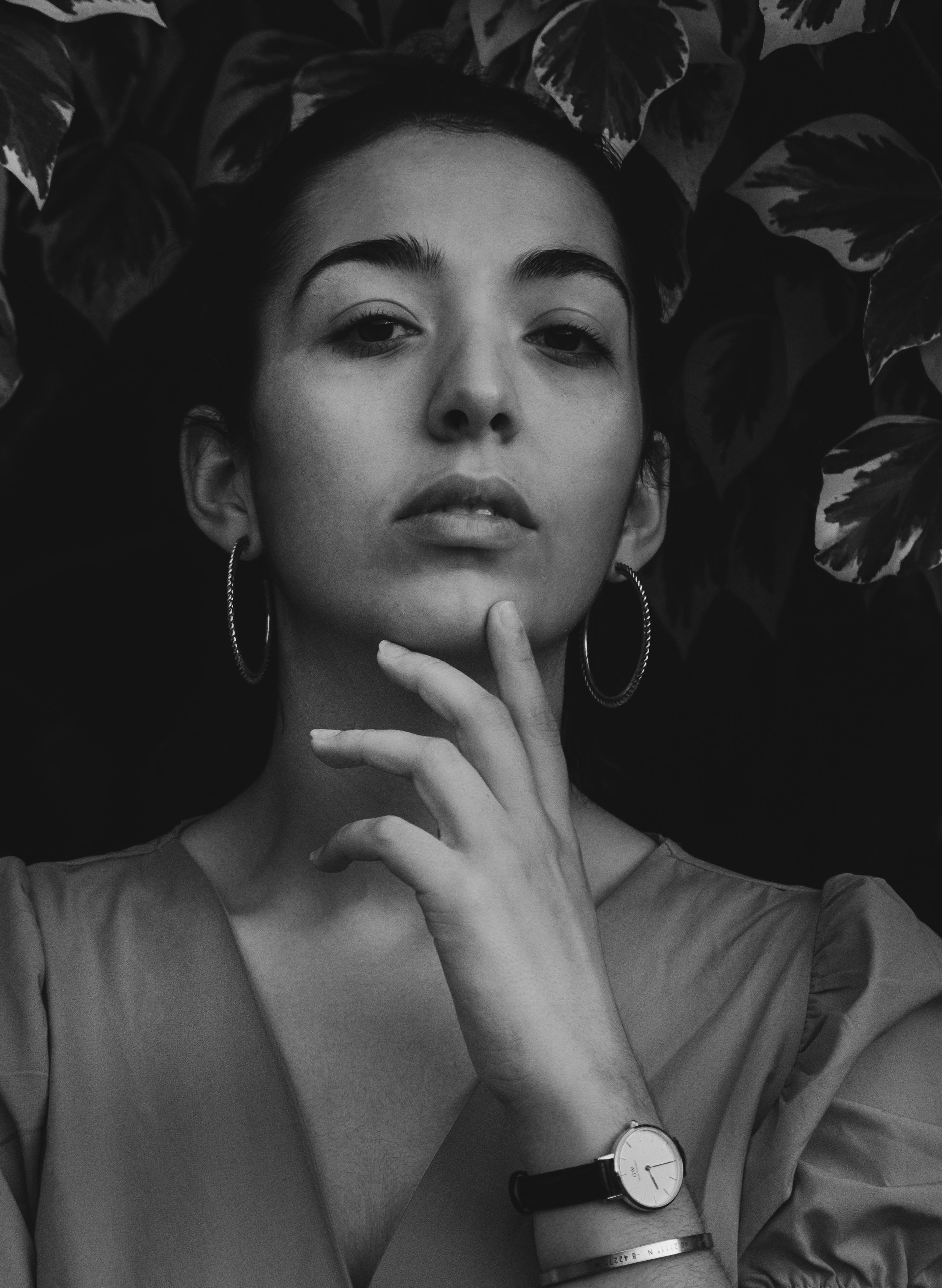 Black and white portrait of a woman with hoop earrings, wearing a watch and bracelet, touching her chin.