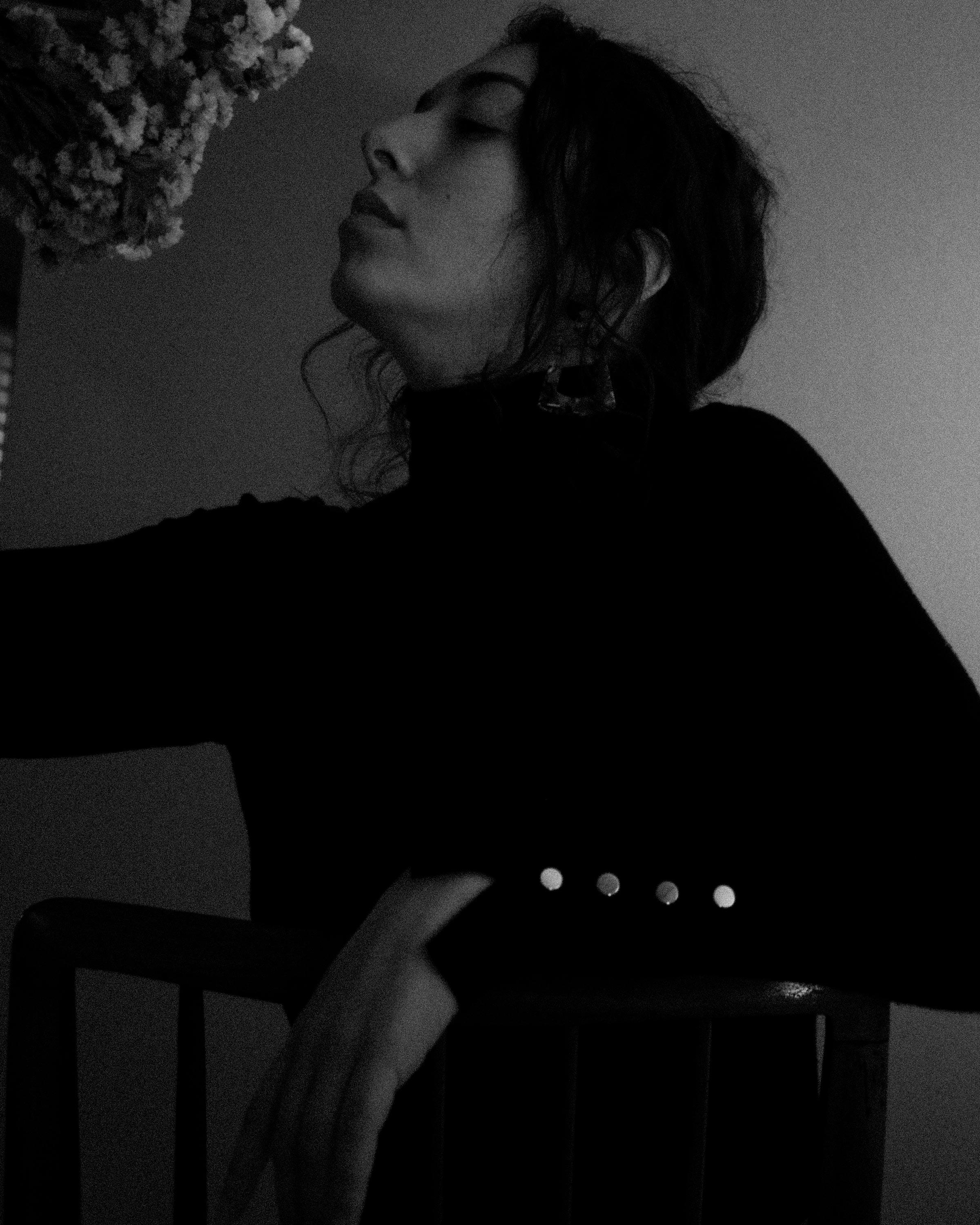 Black and white portrait of a woman in profile wearing a dark turtleneck, resting her arm on a chair while looking at flowers.
