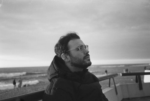 Man with glasses and beard wearing a puffer jacket looking up, sitting near a beach with waves and people in the background.