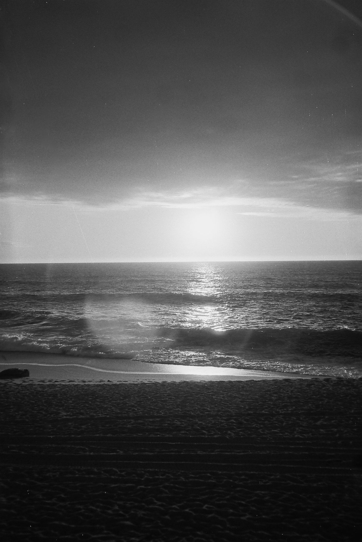 Black and white photo of ocean waves at sunset with the sun low on the horizon and a sandy beach in the foreground.