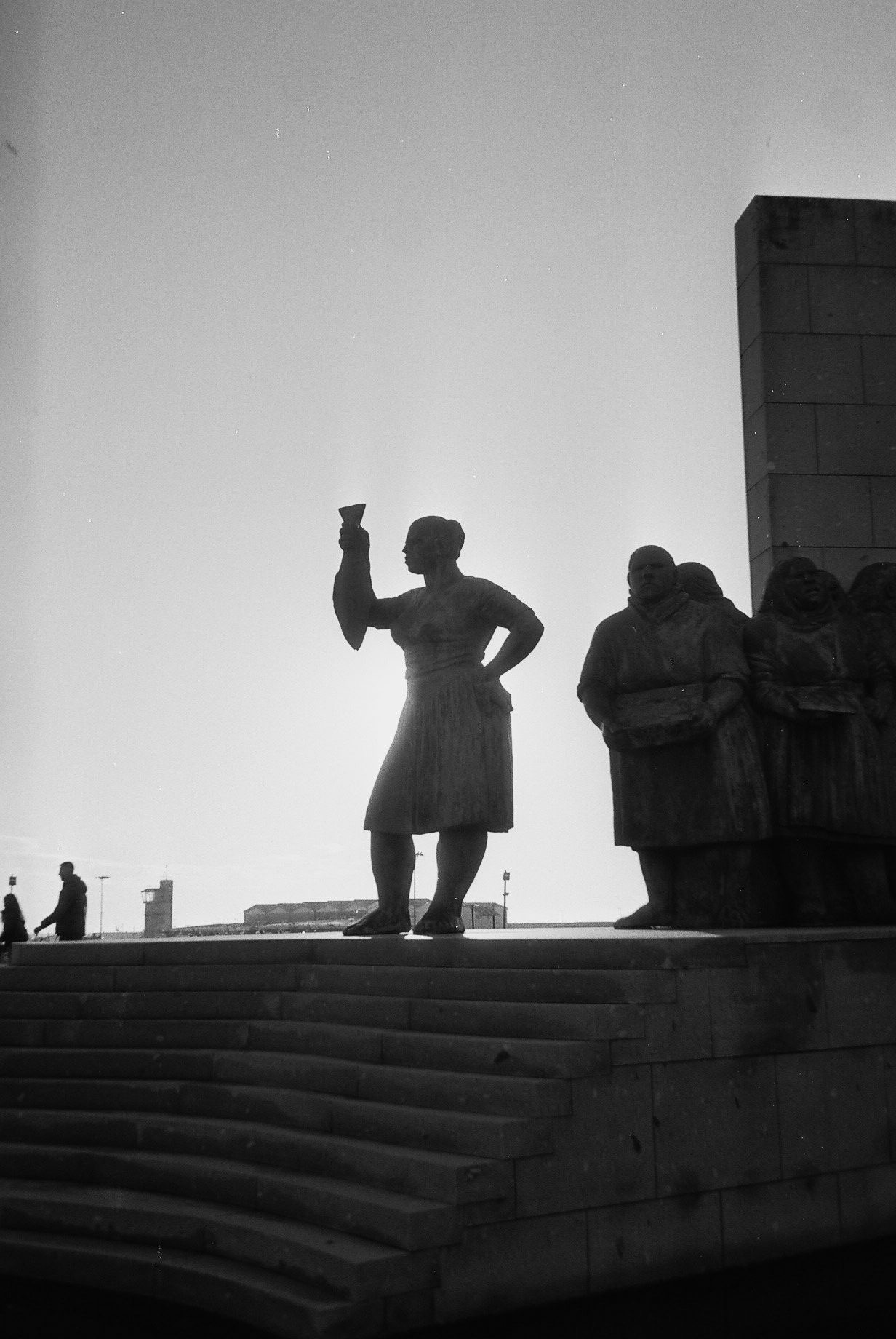 Silhouetted statues of a woman holding a glass and a group of people standing on steps during sunset.
