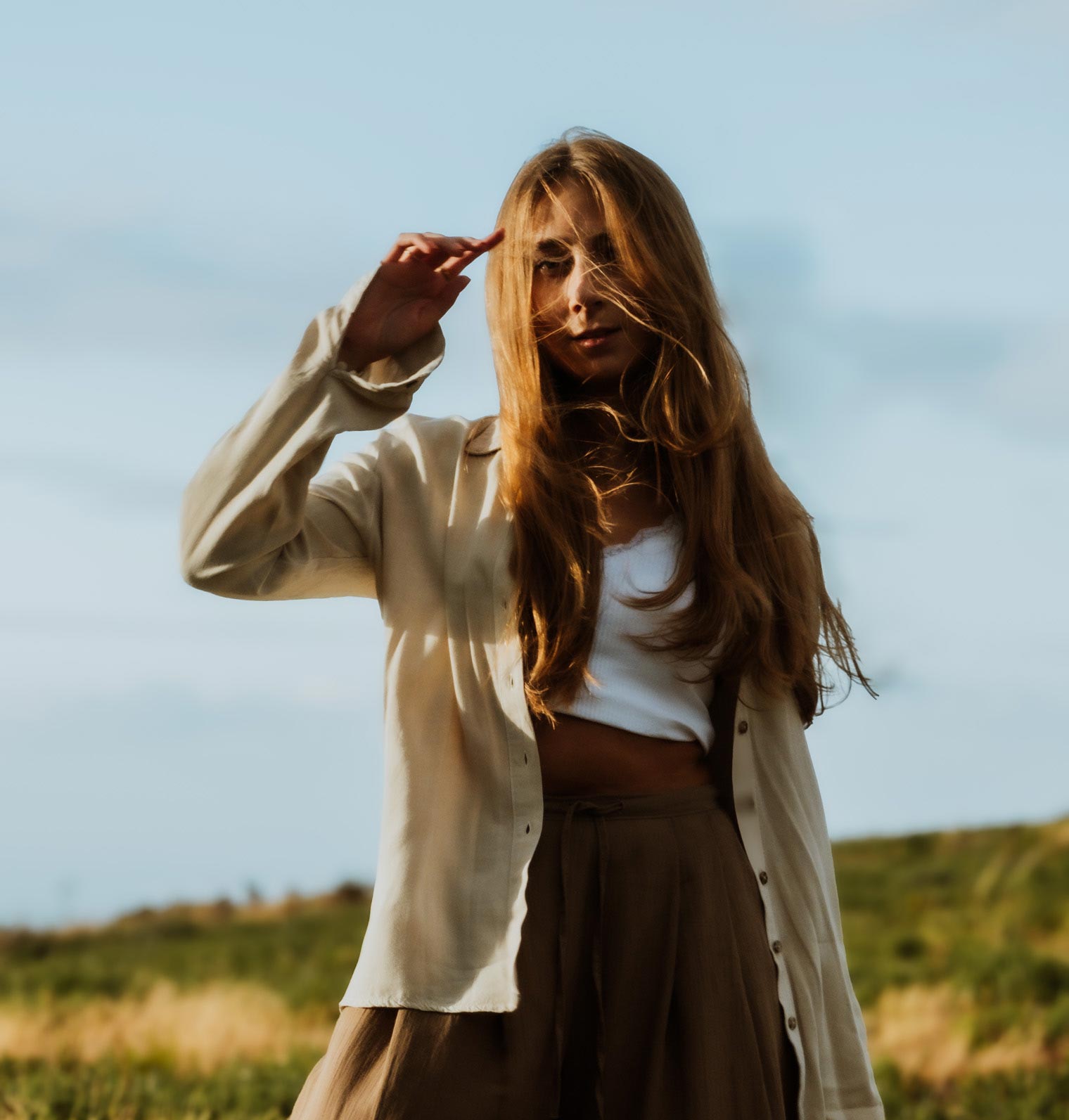 Young woman with long blonde hair wearing a beige shirt and brown pants standing outdoors against a clear sky.