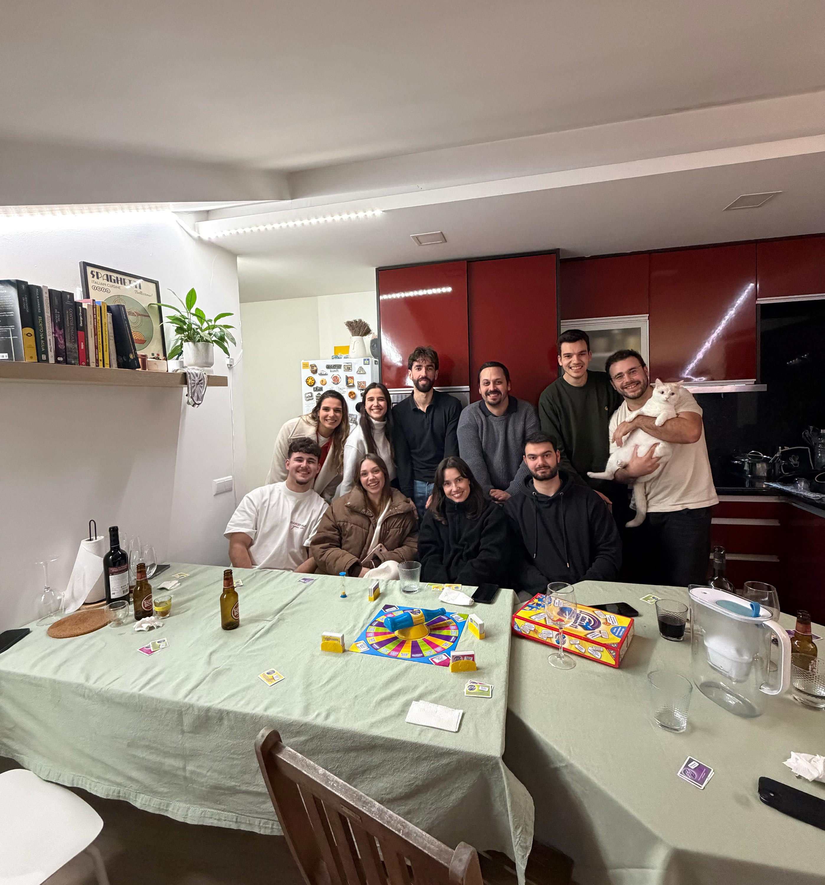 Group of ten young adults gathered in a kitchen around a table playing a board game, one person holding a white cat.
