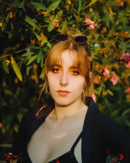 Young woman with light brown hair and sunglasses on her head, standing in front of lush green foliage with pink flowers.
