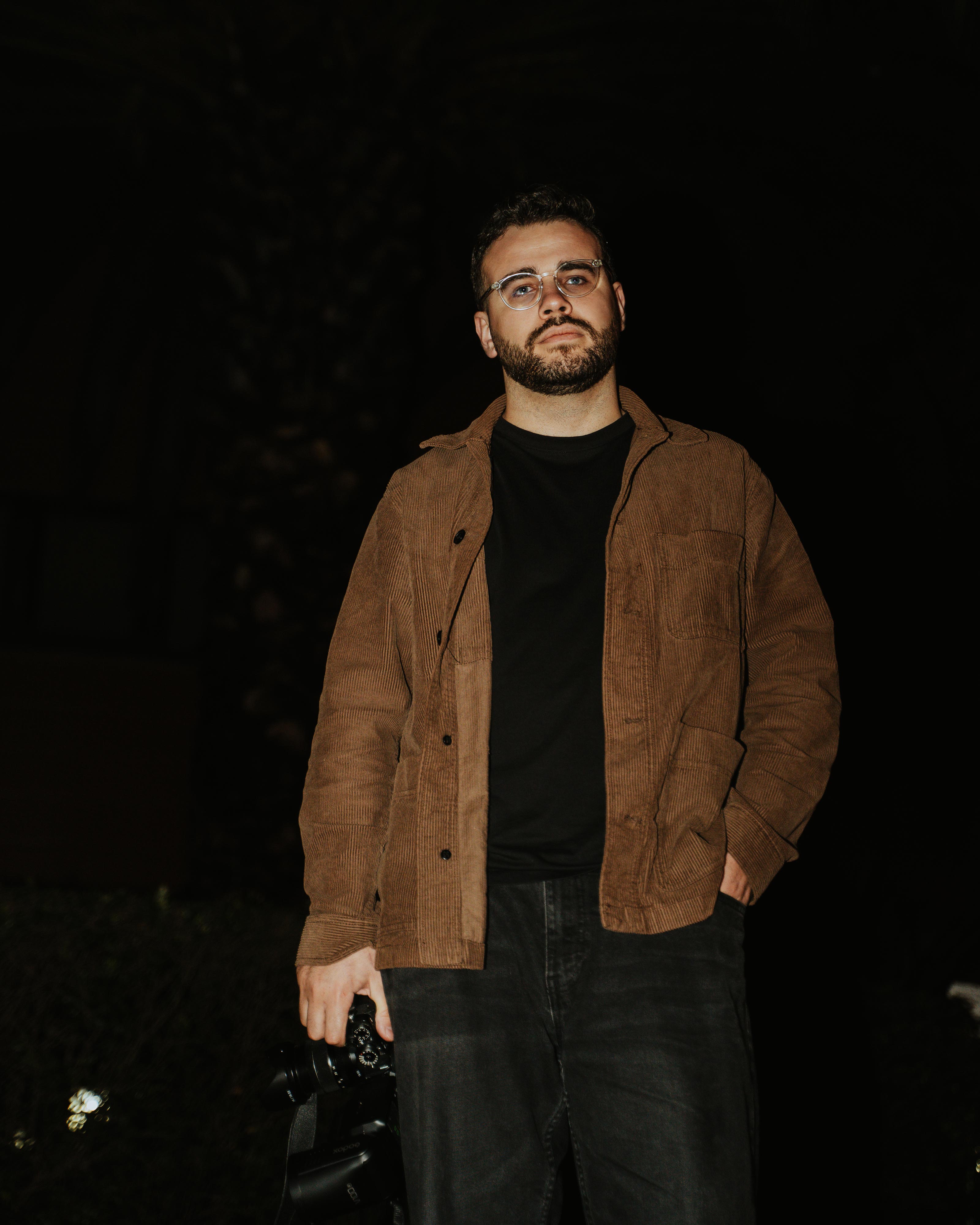 Man with beard and glasses wearing a brown jacket and black shirt holding a camera in one hand at night.