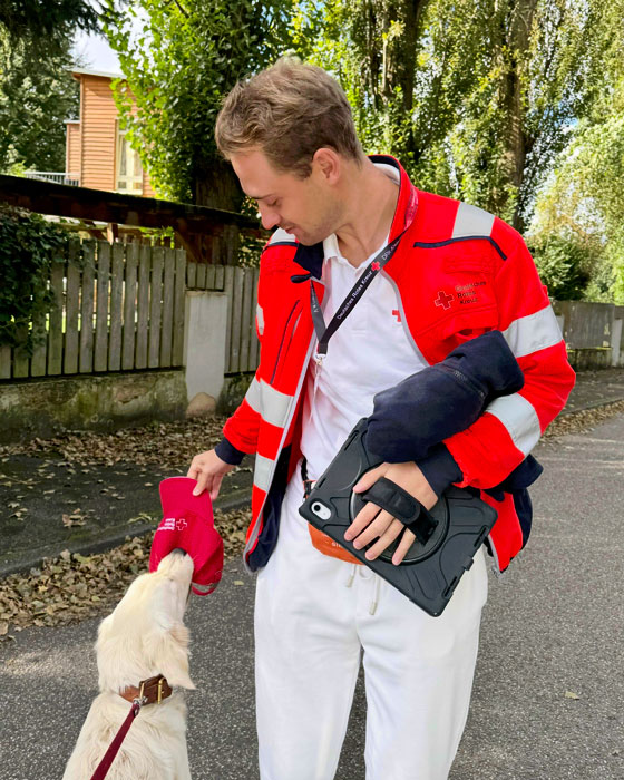 Ein junger Fundraiser in DRK Ausrüstung begegnet einem Golden Retriever auf einer idyllischen Straße.