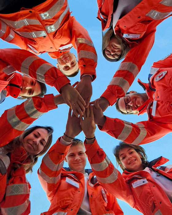 Eine Gruppe von jungen Menschen in der Ausrüstung des Deutschen Roten Kreuzes steht in einem Kreis. Ihre Hände sind ausgestreckt und begegnen sich in der Mitte des Kreises. 