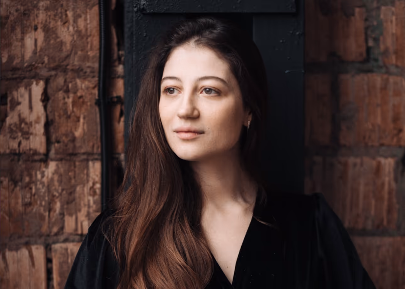 Жінка з довгим каштановим волоссям у чорному вбранні стоїть біля цегляної стіни, дивлячись убік.