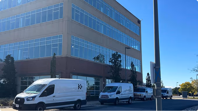 Four white commercial vans parked on the street beside a modern multi-story office building under a clear blue sky.