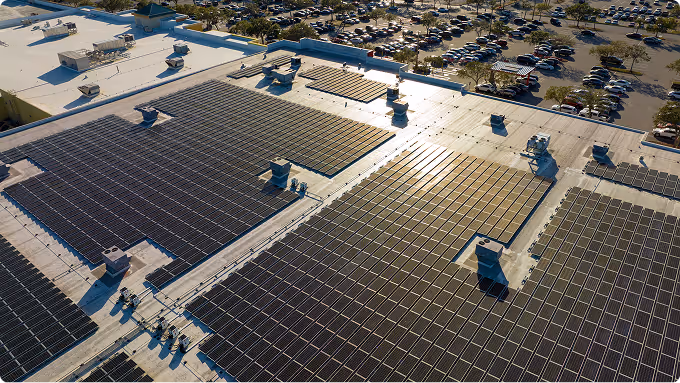 Aerial view of large solar panel arrays installed on a flat rooftop with a busy parking lot in the background.