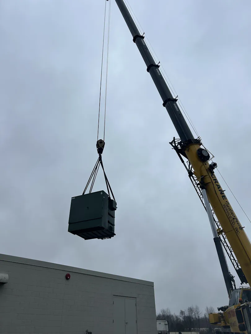 Yellow crane lifting a large green industrial generator or HVAC unit above a building on a cloudy day.