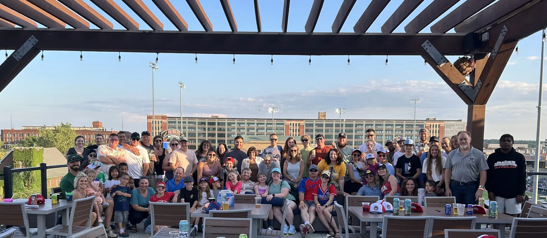 Large group of people of various ages gathered under wooden pergola with drinks, smiling and posing for a photo at an outdoor venue with a city building in the background.
