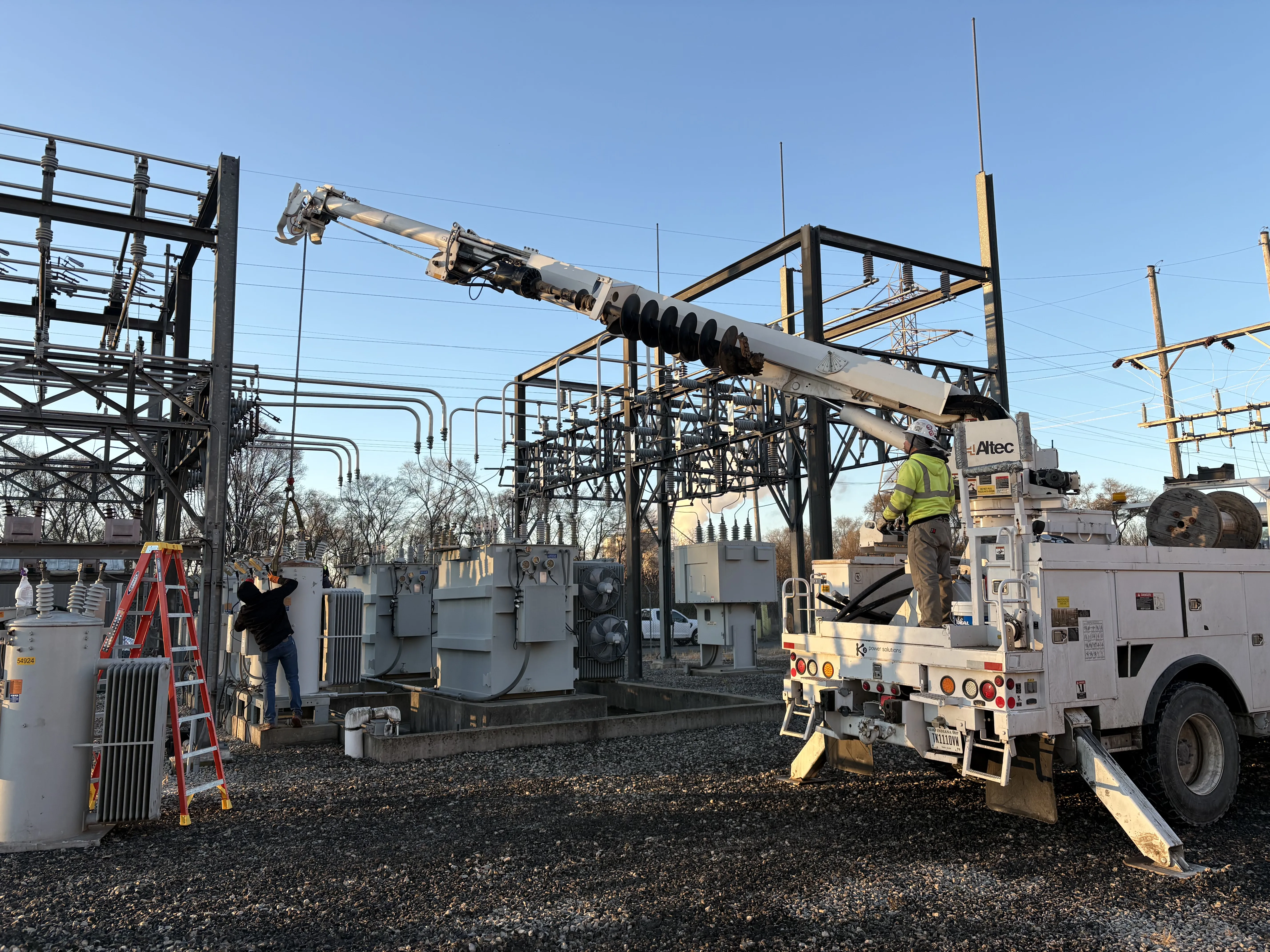 Workers operating a bucket truck to maintain electrical transformers and power lines in an outdoor substation under clear blue sky.