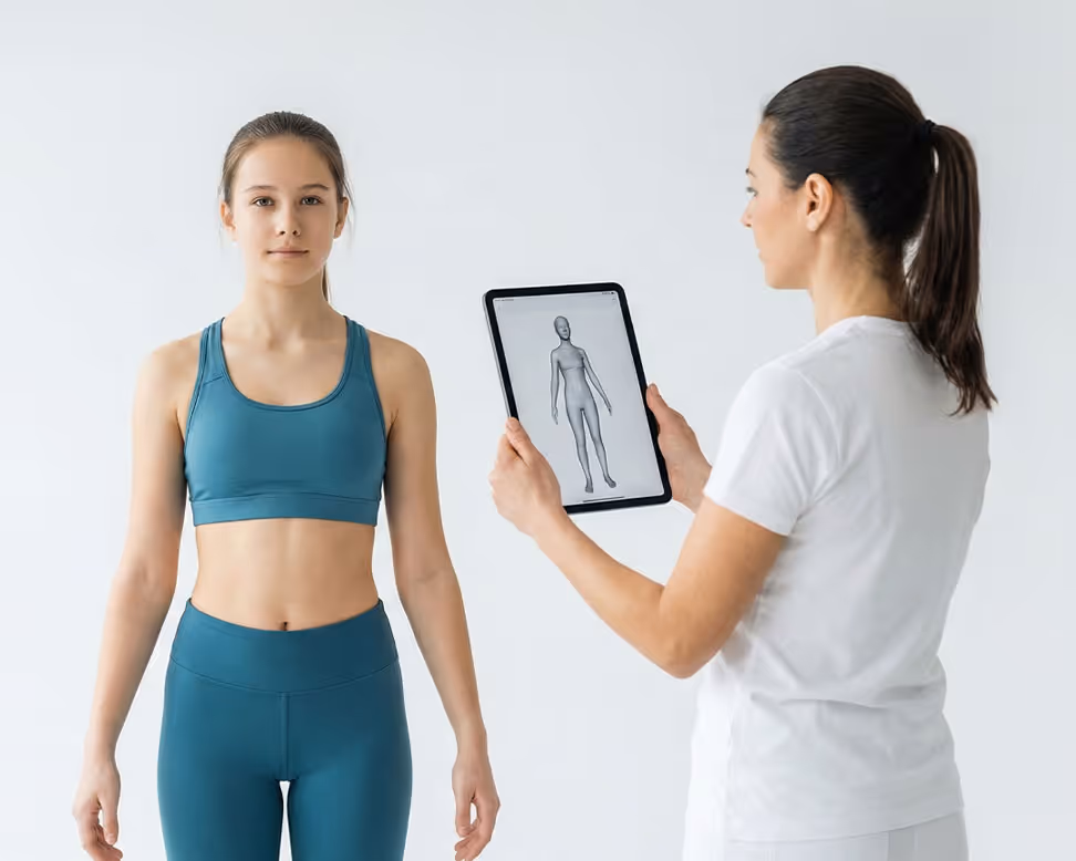 Woman in white shirt holding tablet showing a 3D human body model while a young woman in athletic wear stands facing forward.