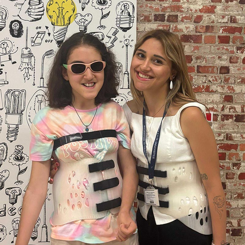 Two smiling women wearing white back braces, one in sunglasses and a tie-dye shirt, the other with a lanyard, standing in front of a brick wall and a wall with lightbulb doodles.