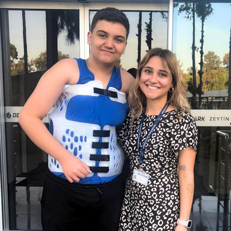 A young man wearing a blue and white medical brace standing next to a smiling woman in a black and white patterned dress with a lanyard and watch, both posing outdoors in front of glass doors.