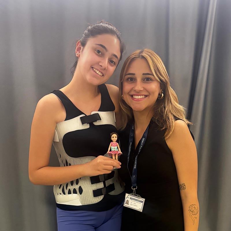 Two smiling women pose closely together; one woman wears a medical back brace and holds a small doll, the other wears a black sleeveless dress with a name badge and has tattoos on her arm.
