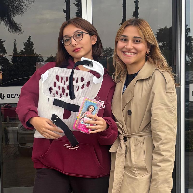 Two young women standing outside a glass door; the woman on the left wearing glasses and a maroon hoodie holds a back brace and a doll action figure, while the smiling woman on the right wears a beige trench coat.