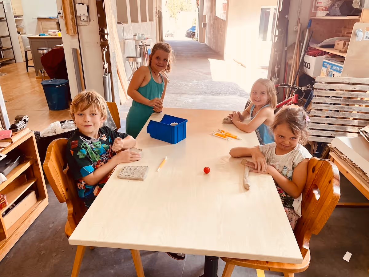 Four children sitting around a table in a workshop, shaping clay with tools and smiling at the camera.