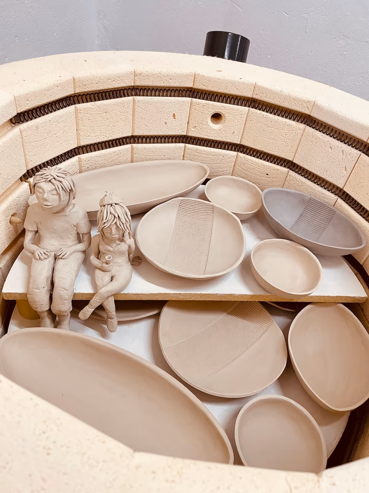 Ceramic kiln interior with unfired beige pottery plates and two small clay figurines sitting on a shelf.