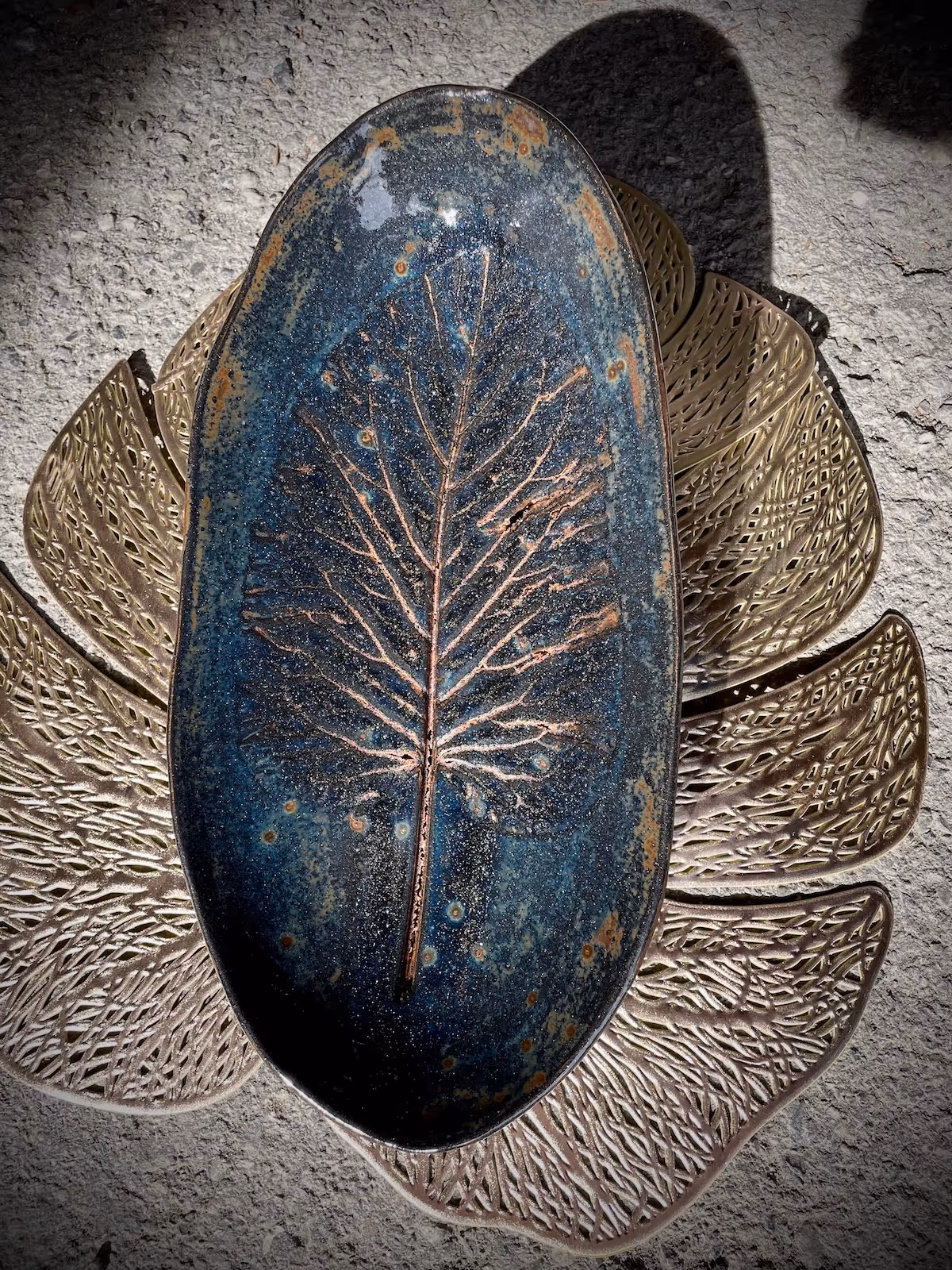 Decorative dark blue ceramic dish with a raised leaf pattern, placed on a metallic leaf-shaped tray on textured ground.