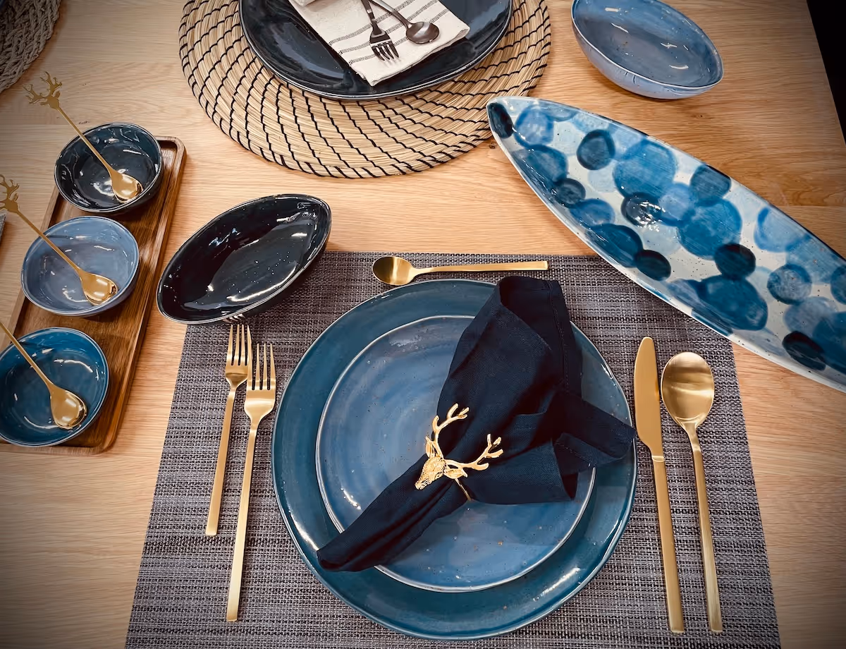 Table setting with blue ceramic plates, gold cutlery, black cloth napkin with deer-shaped gold napkin ring, and small blue bowls on a wooden table.