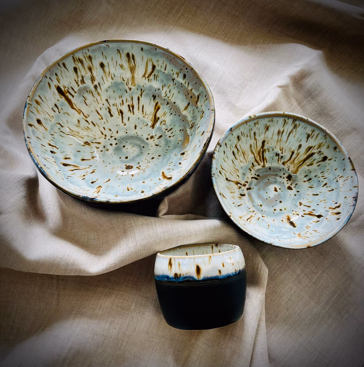 Three ceramic dishes with speckled brown and blue glaze resting on beige fabric.
