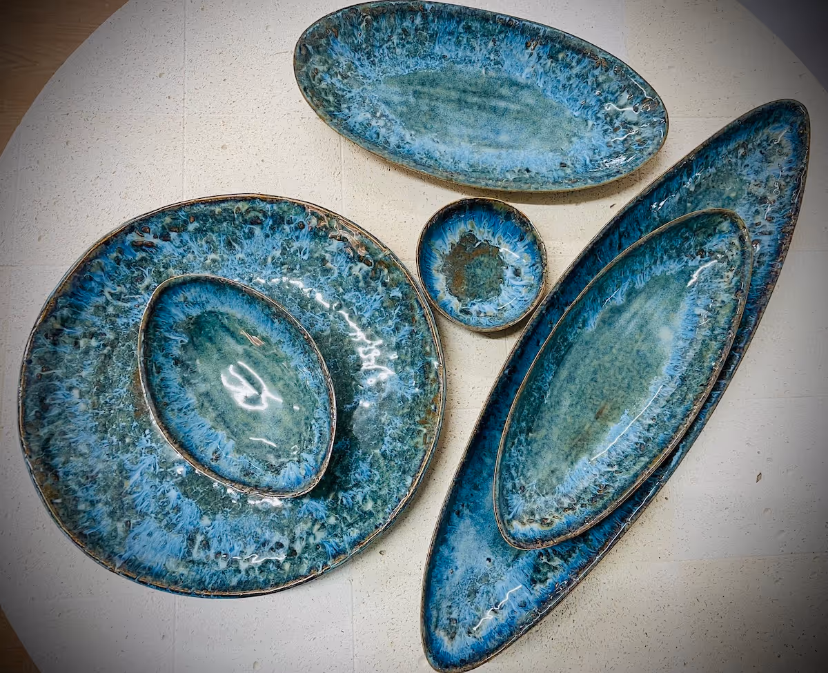 Set of ceramic dishes in varying oval and round shapes with a blue-green mottled glaze, arranged on a white textured surface.