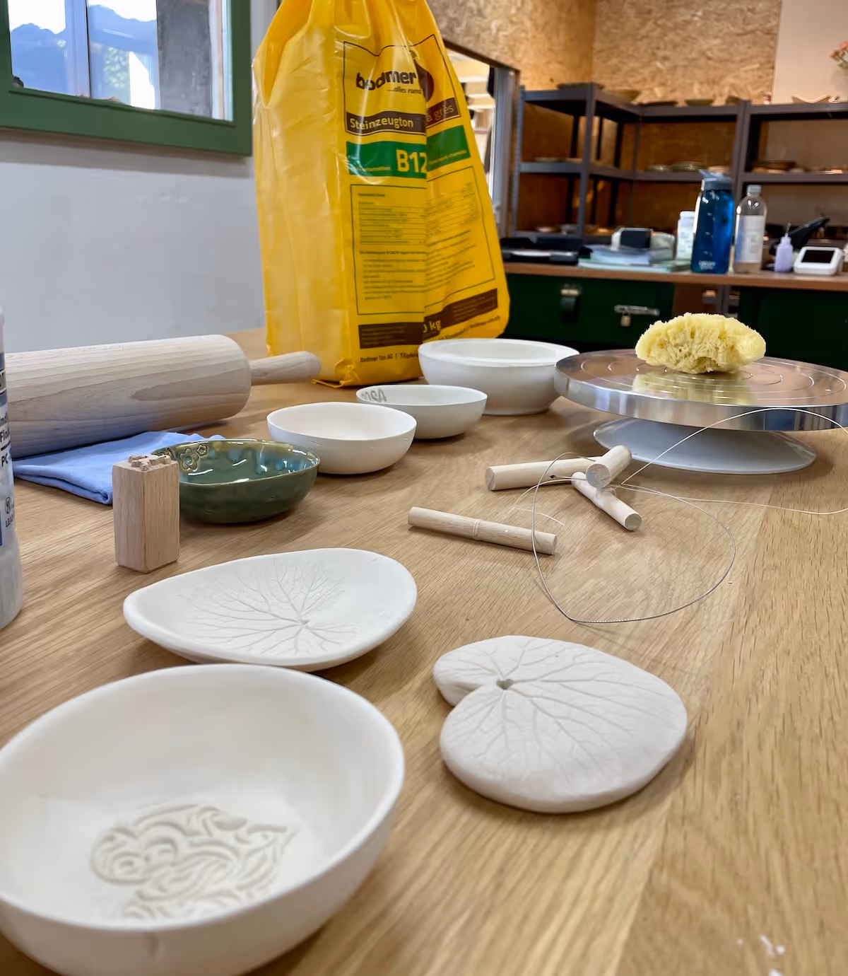 Wooden table with ceramic bowls and leaf-shaped ceramic pieces, a rolling pin, pottery tools, a yellow bag of clay, and a sponge in a pottery workshop.