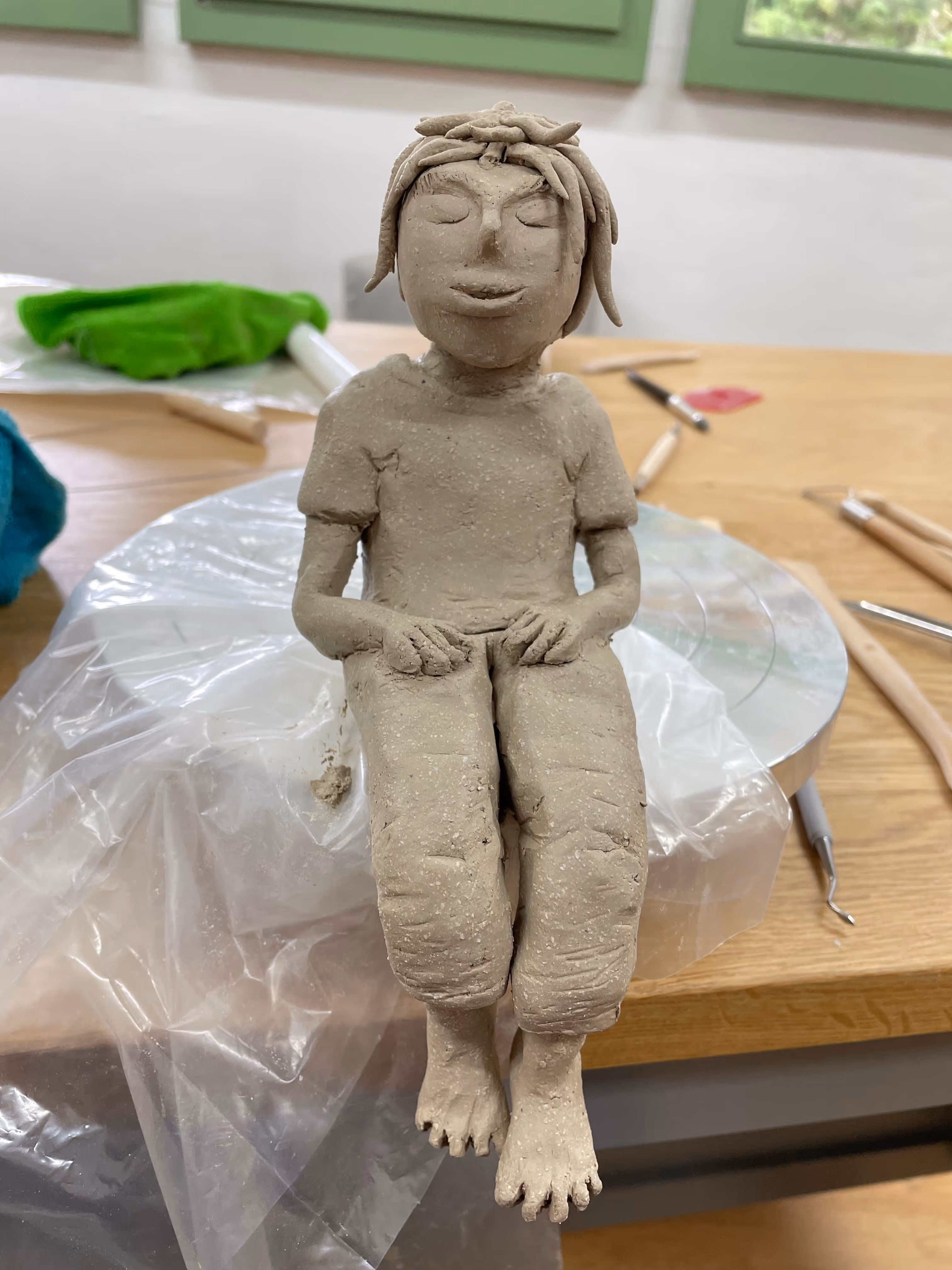 Handmade clay sculpture of a seated person with closed eyes and textured hair, placed on a turntable covered with plastic on a wooden table.