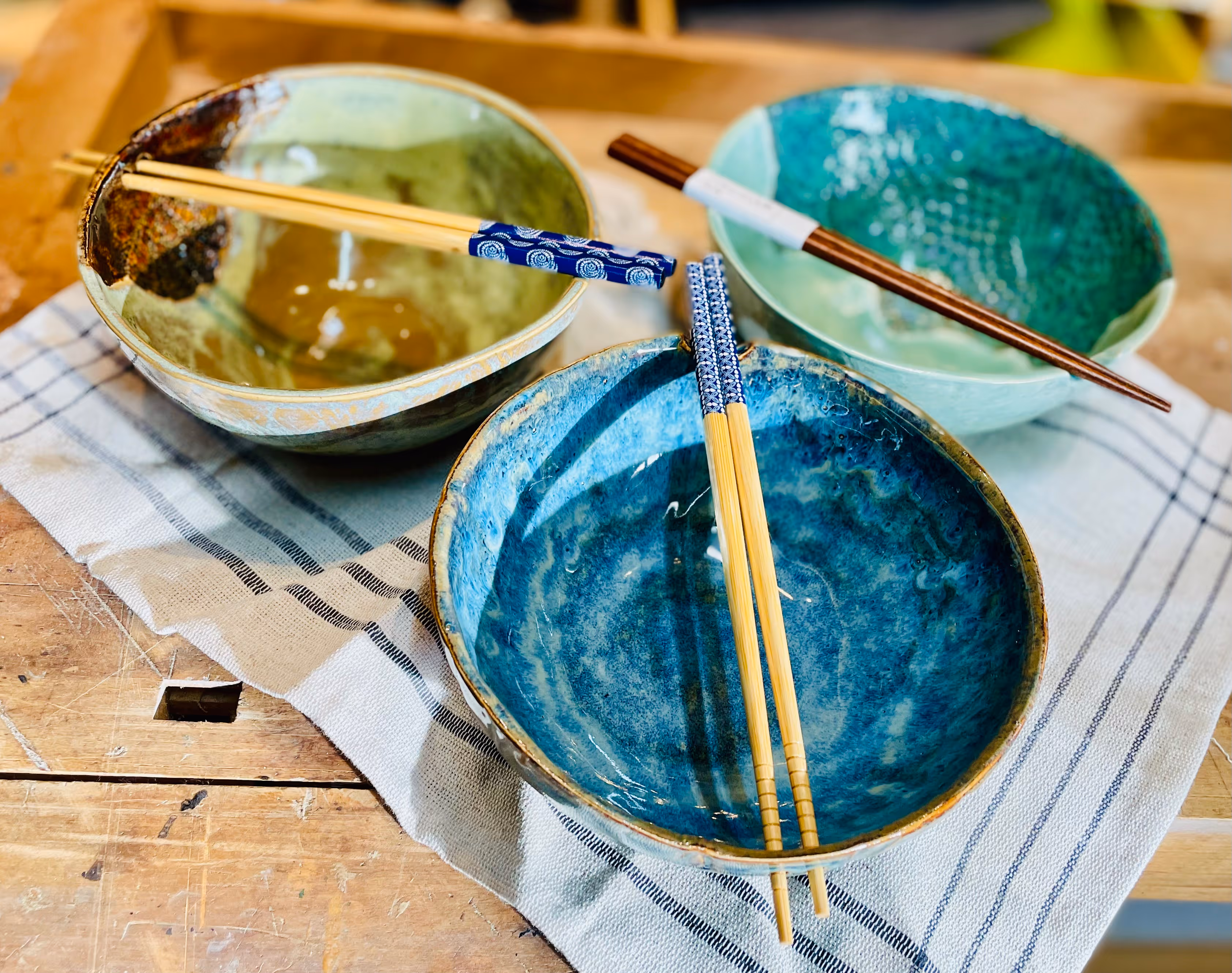 Three ceramic bowls in green and blue shades with chopsticks resting on each, placed on a striped cloth on a wooden surface.