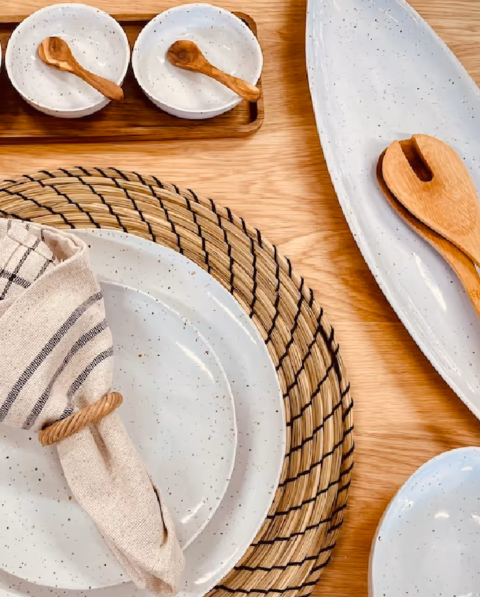 Table setting with speckled white ceramic plates, a woven placemat, beige striped napkin with ring, two small bowls with wooden spoons, and an elongated dish with wooden salad servers on a wooden surface.